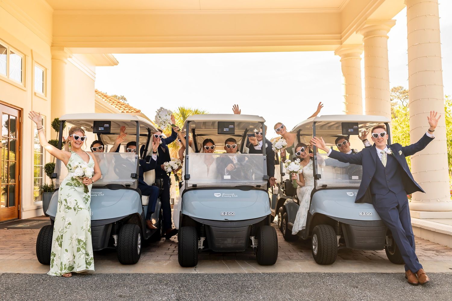 Wedding party celebrates joyfully while posing with golf carts outside a grand venue entrance.