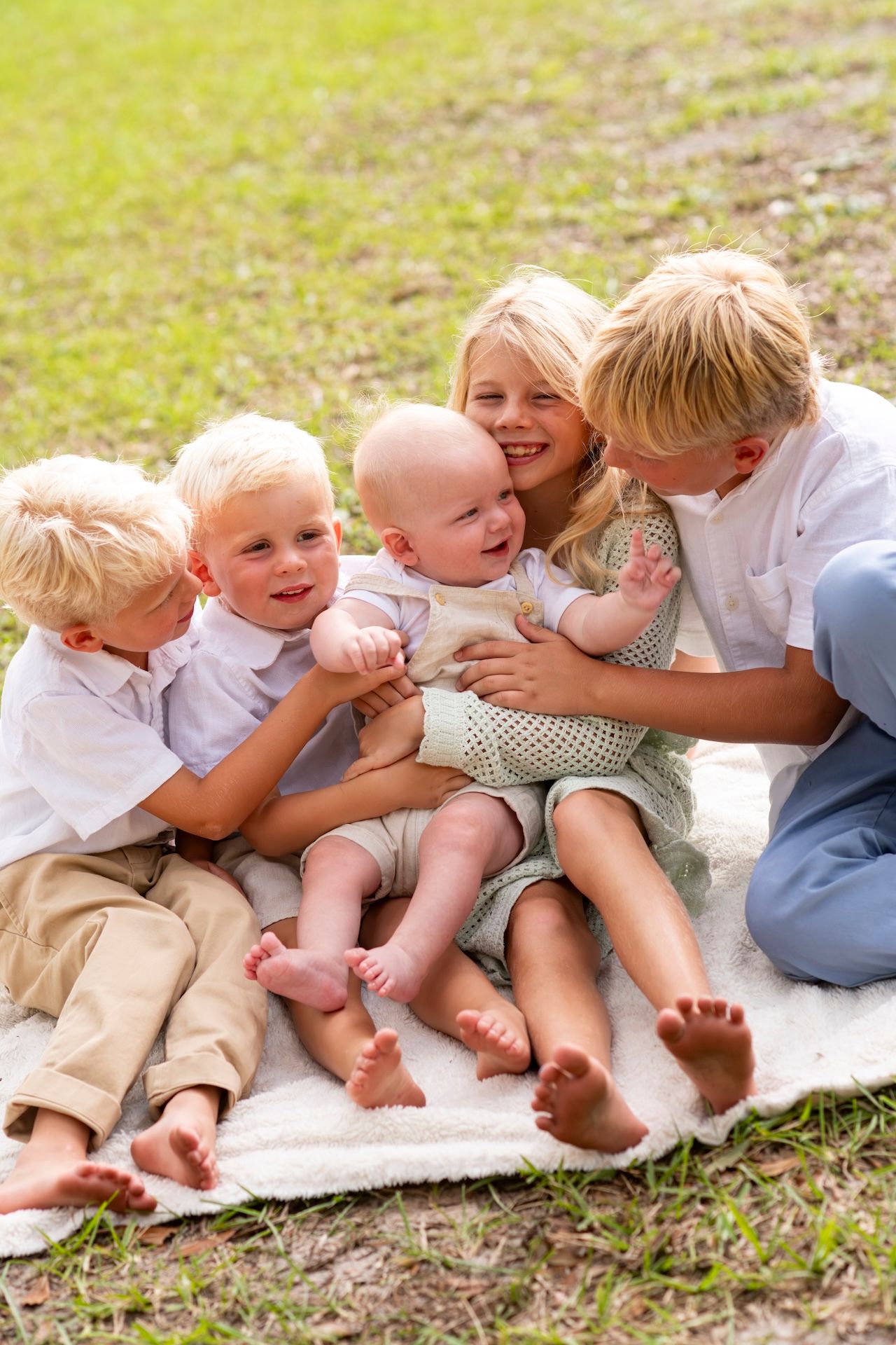 Five young blonde siblings sit together on a blanket outdoors, hugging a baby in a sunny green park.