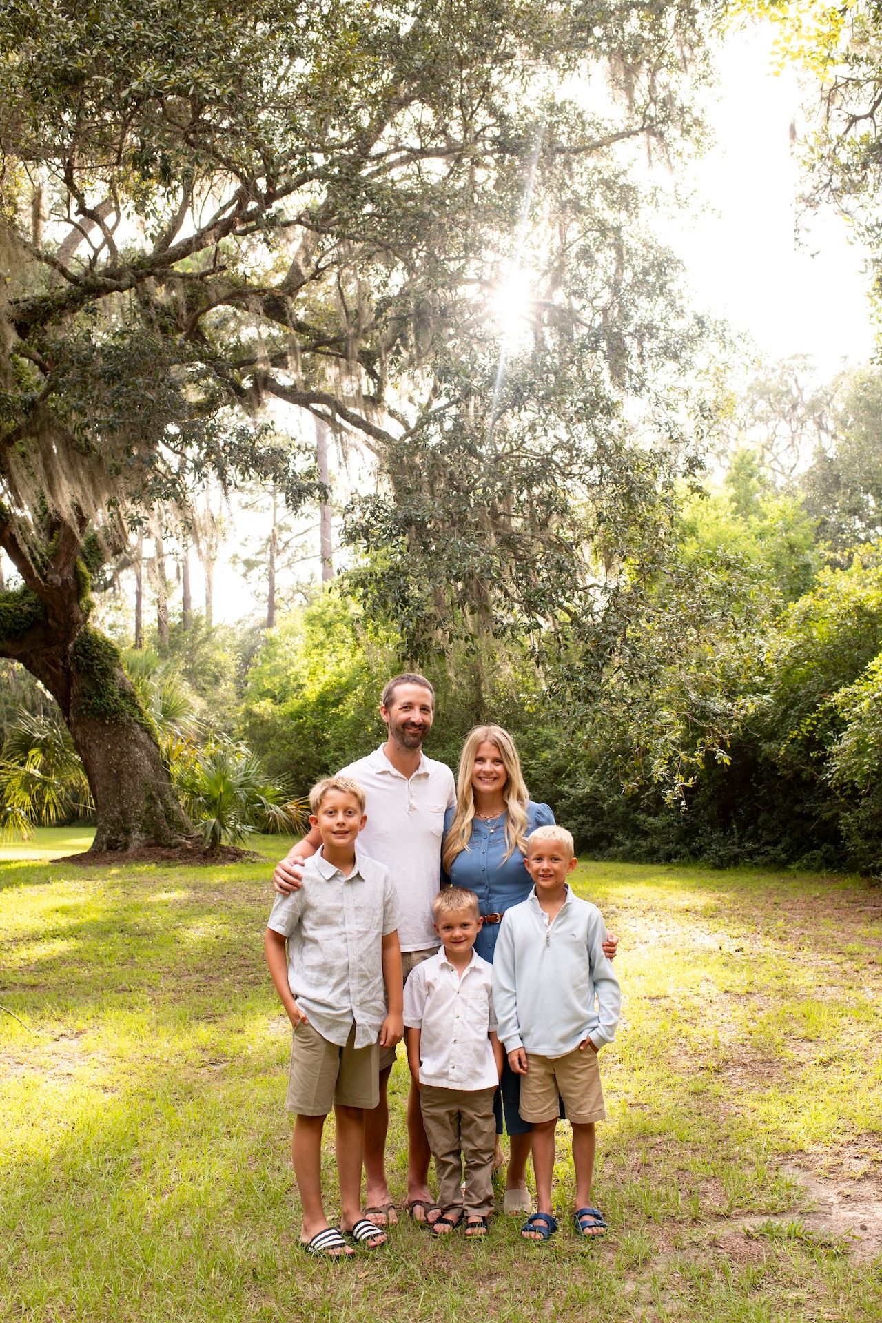 Family of five posing outdoors under a large oak tree with Spanish moss in a lush green park setting.