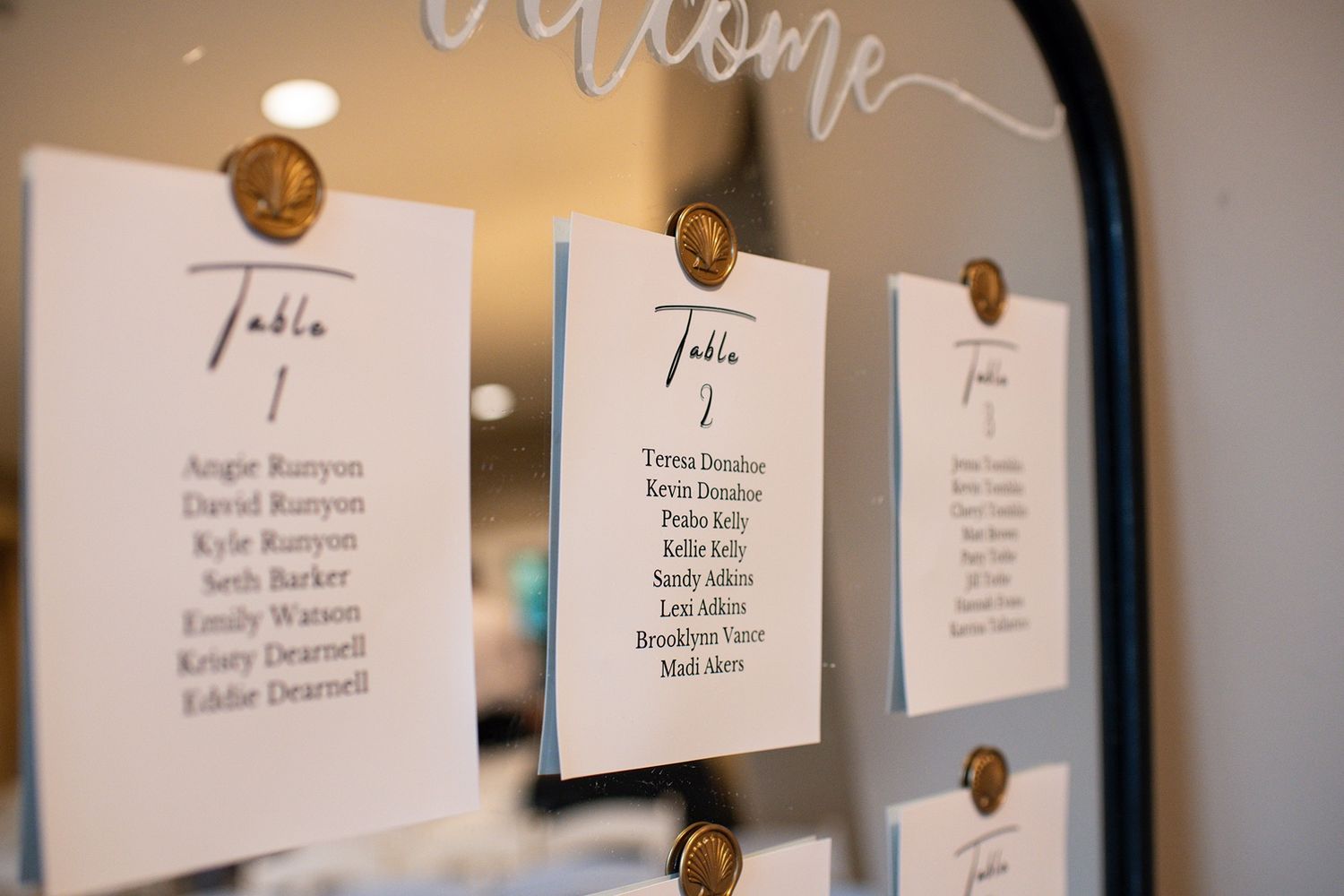 Wedding seating chart cards displayed on a wall with elegant gold pins and calligraphy text.