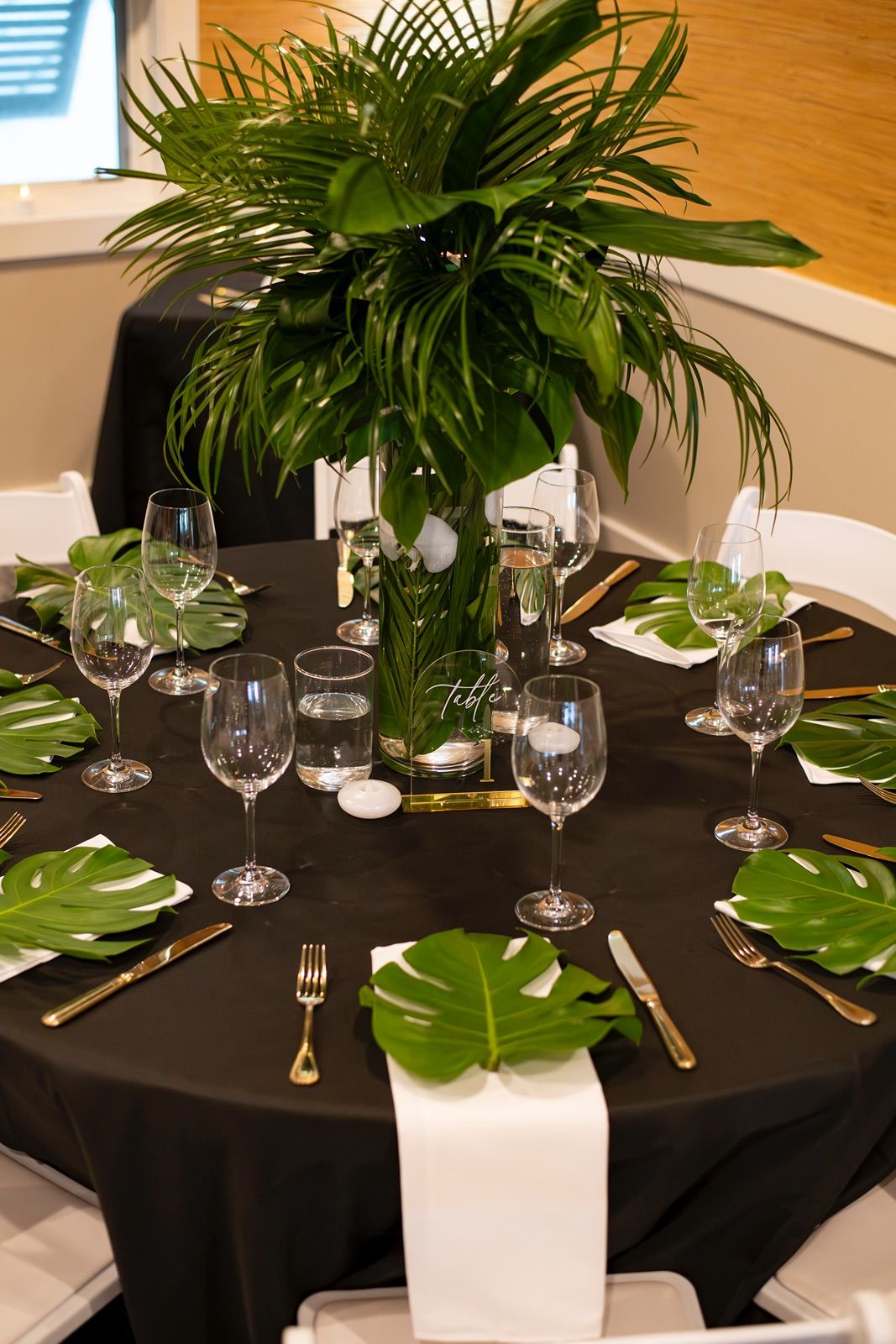 Elegant table settings with black tablecloths, tropical palm leaves, and wine glasses create a sophisticated dining atmosphere.