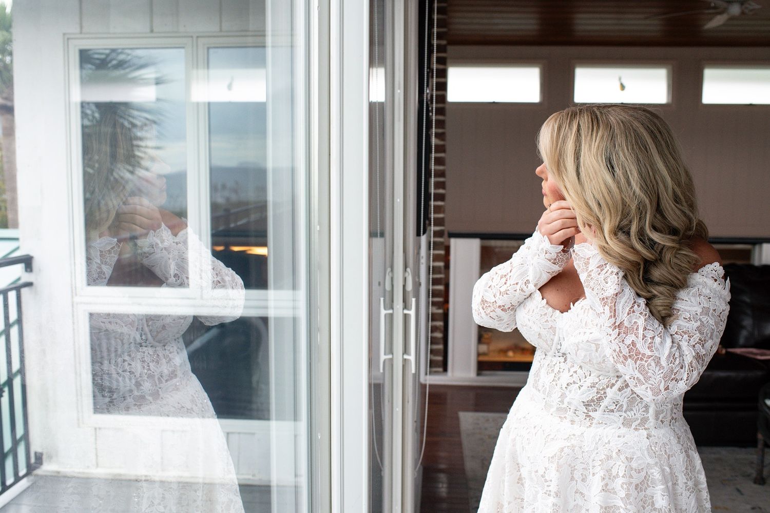 A bride in a long-sleeved lace wedding dress puts on earrings while getting ready near a window.