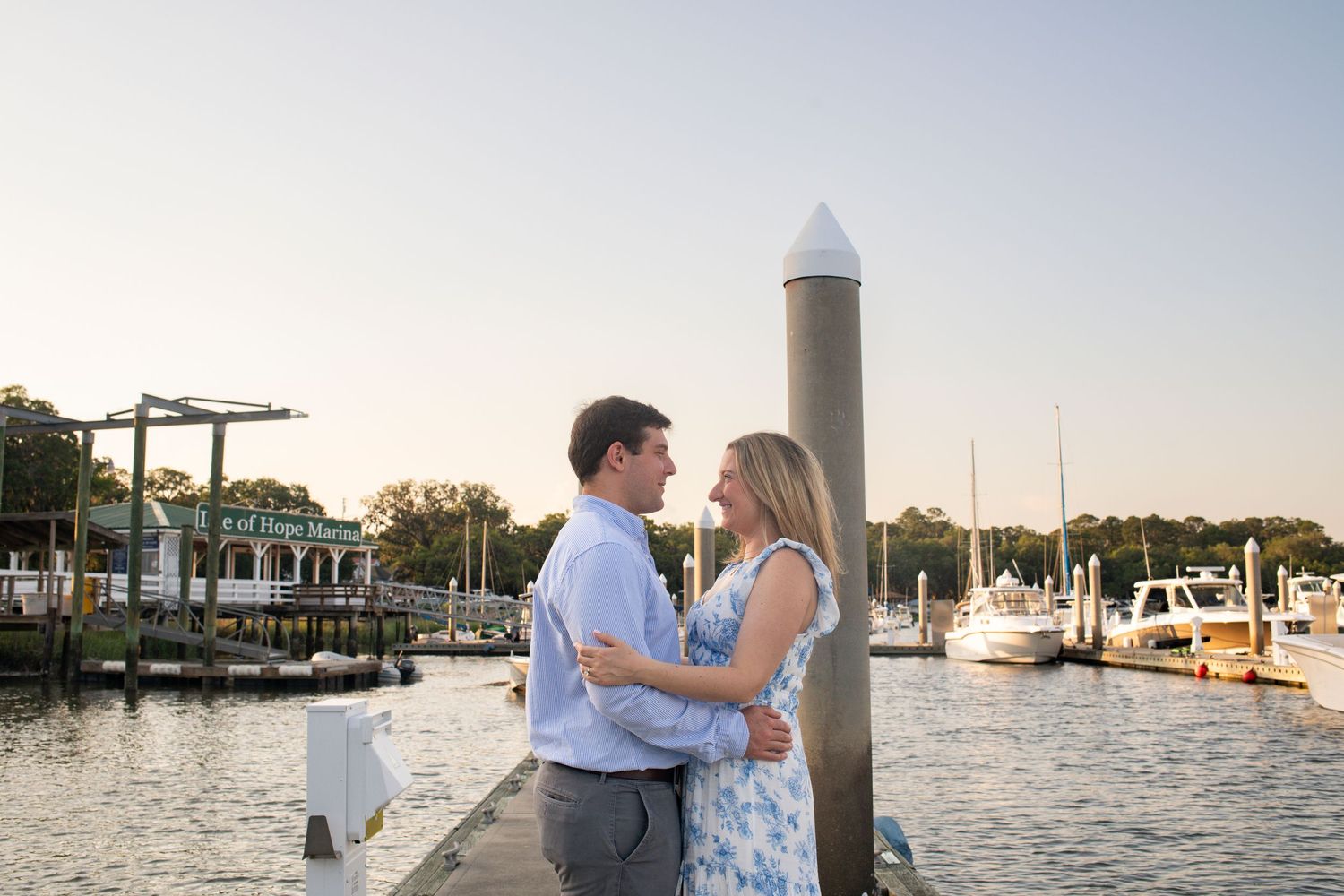 A couple shares a romantic moment at sunset by the marina with boats and water in the background.