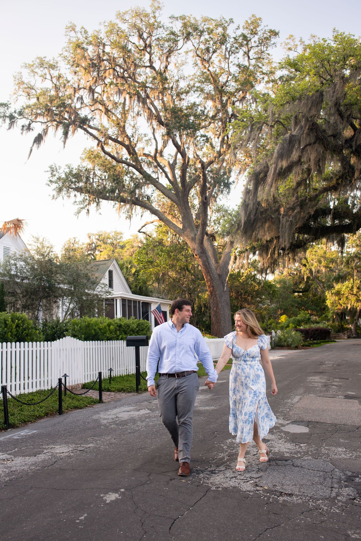 A couple walks together on a tree-lined path with Spanish moss and a white picket fence in the background.