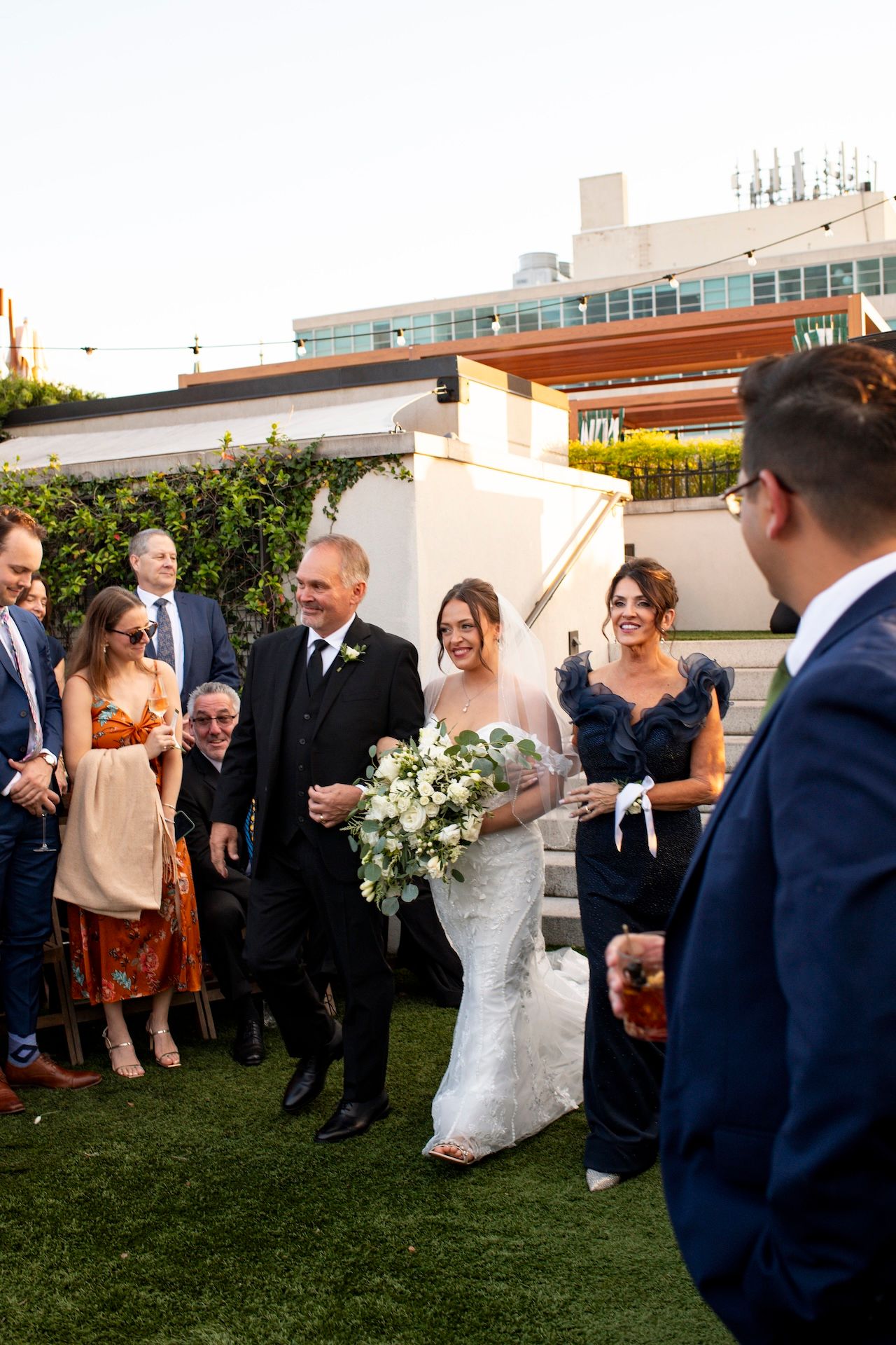 Wedding guests gather on a grassy rooftop venue during an outdoor celebration at sunset.