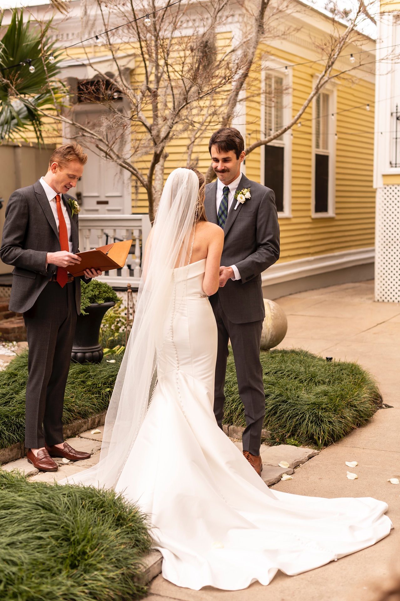 Wedding ceremony takes place in front of yellow historic building with bride in flowing white dress.