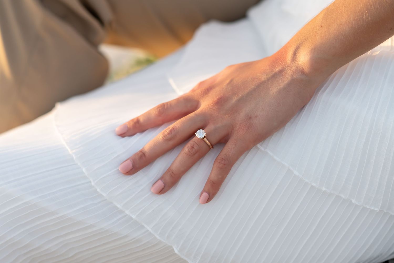 Close-up of a hand wearing an engagement ring resting on white bedding.