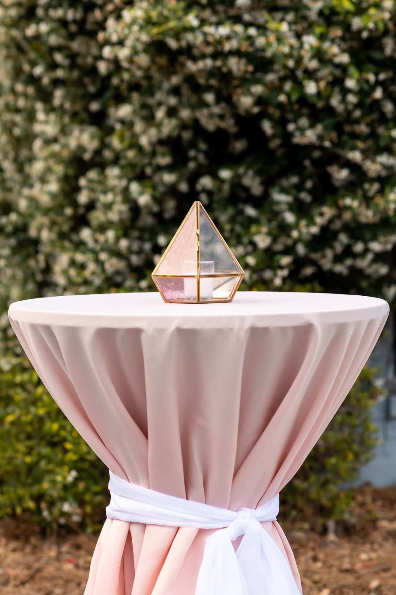 Light pink linens drape elegantly over circular cocktail tables at an outdoor event venue with lush greenery in the background.