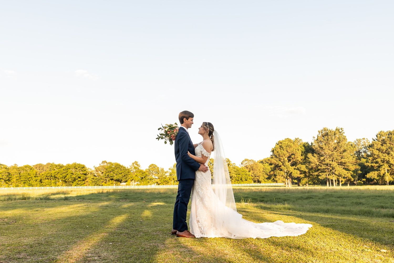 Couple embraces in golden sunlight on grassy field during outdoor wedding ceremony in late afternoon.