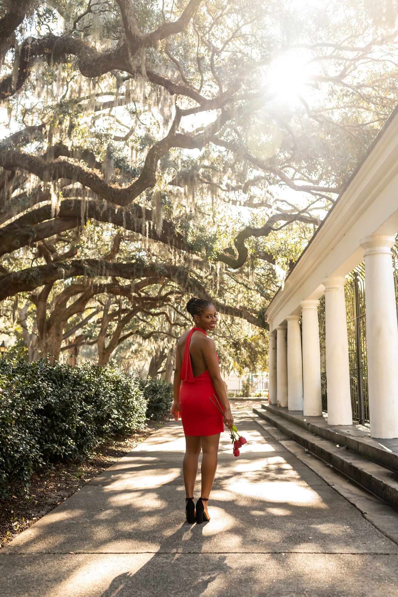 A figure in a vibrant red dress stands on a walkway beneath towering oak trees draped with Spanish moss in the sunlight.