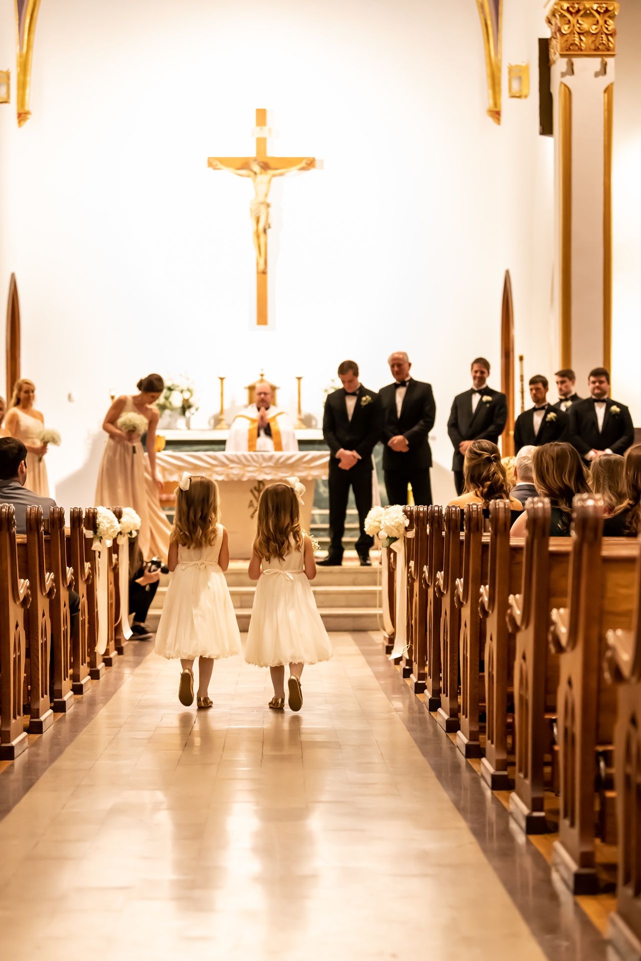 Two flower girls in matching white dresses walk down the aisle during a traditional Catholic church wedding ceremony.