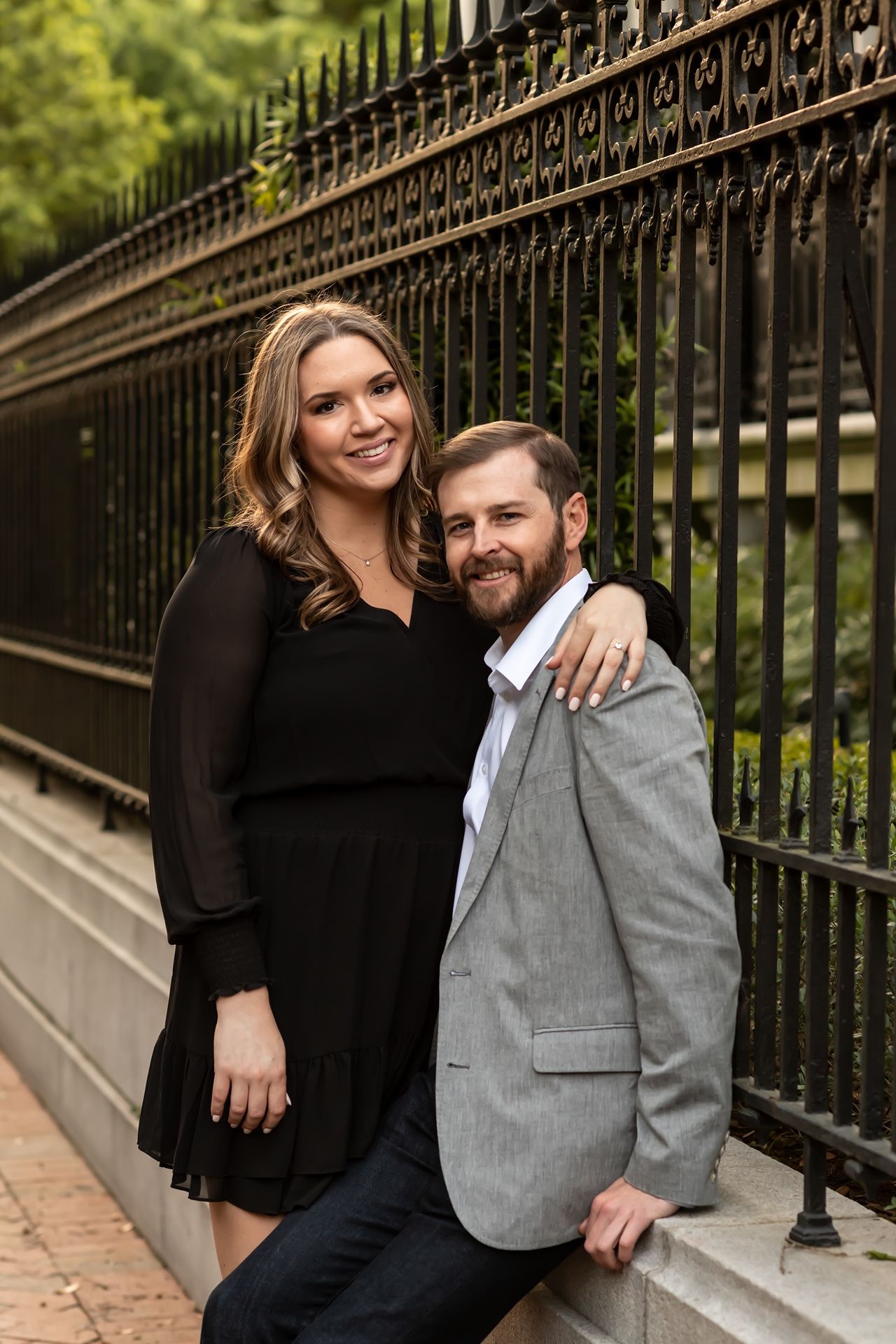 A couple poses together by a decorative iron fence, with the person in black embracing the one in a gray blazer.