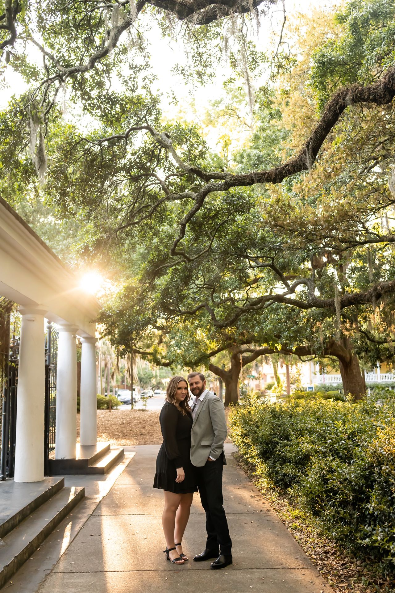 A romantic couple shares tender moments along a sunlit pathway lined with oak trees and white columns.