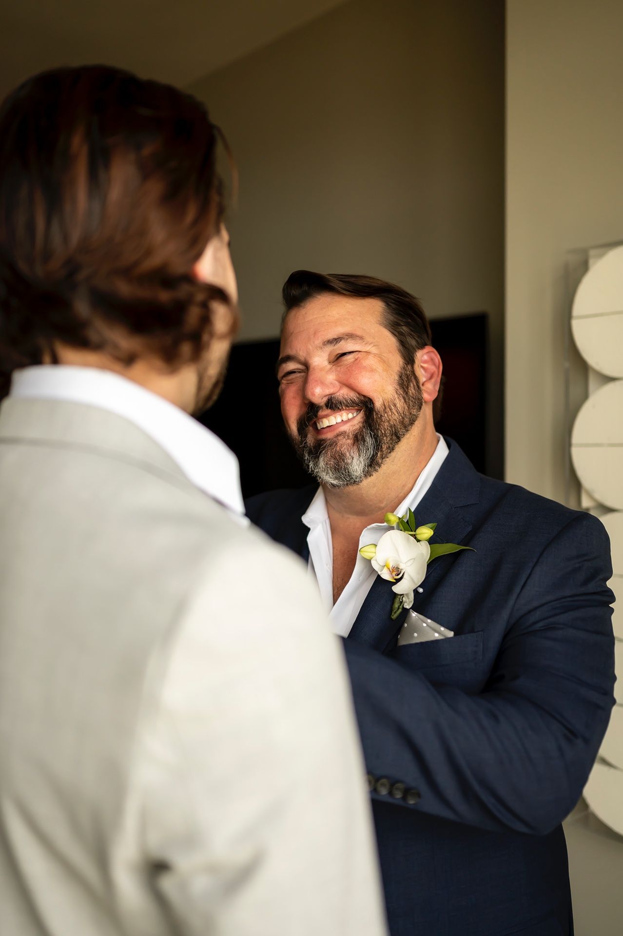 A person in a navy suit with a beard and white boutonniere smiles warmly during what appears to be a wedding moment.