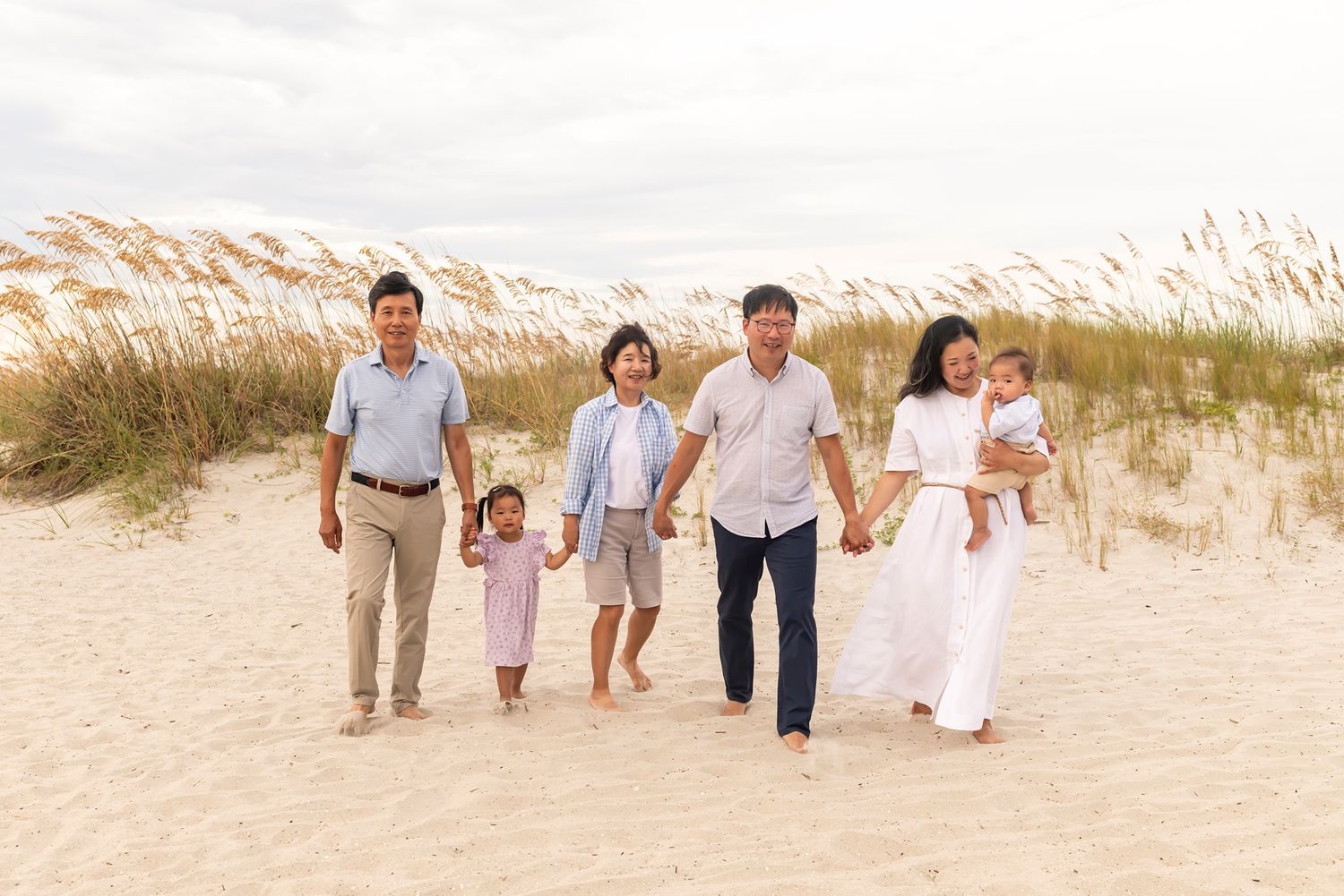 A group wearing white and beige outfits walks together along a sandy beach with sea grass in the background.