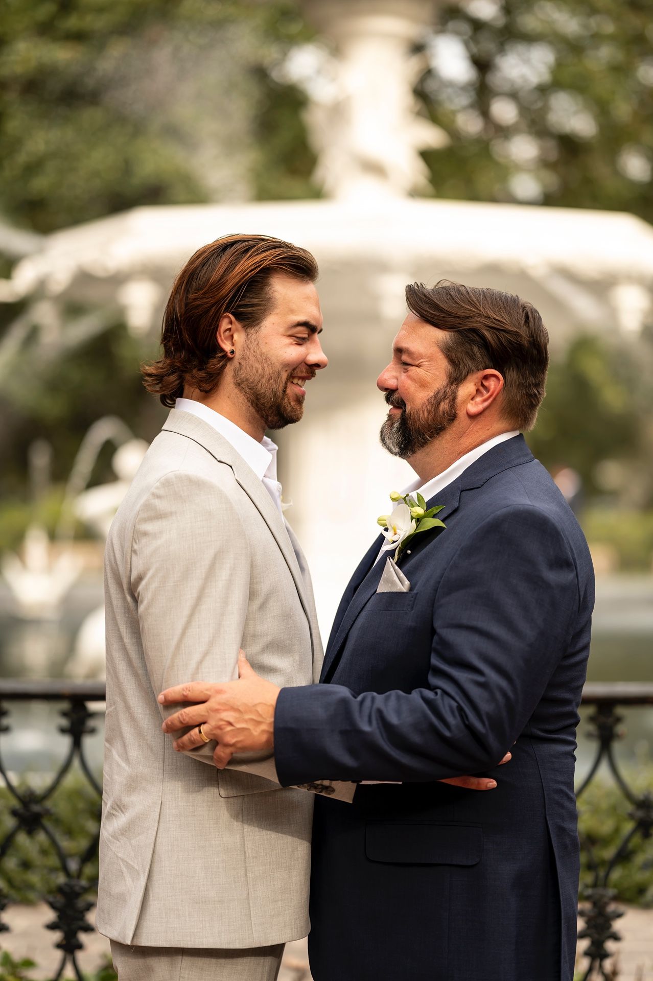 Two grooms share an intimate moment in front of a garden fountain during their wedding celebration.