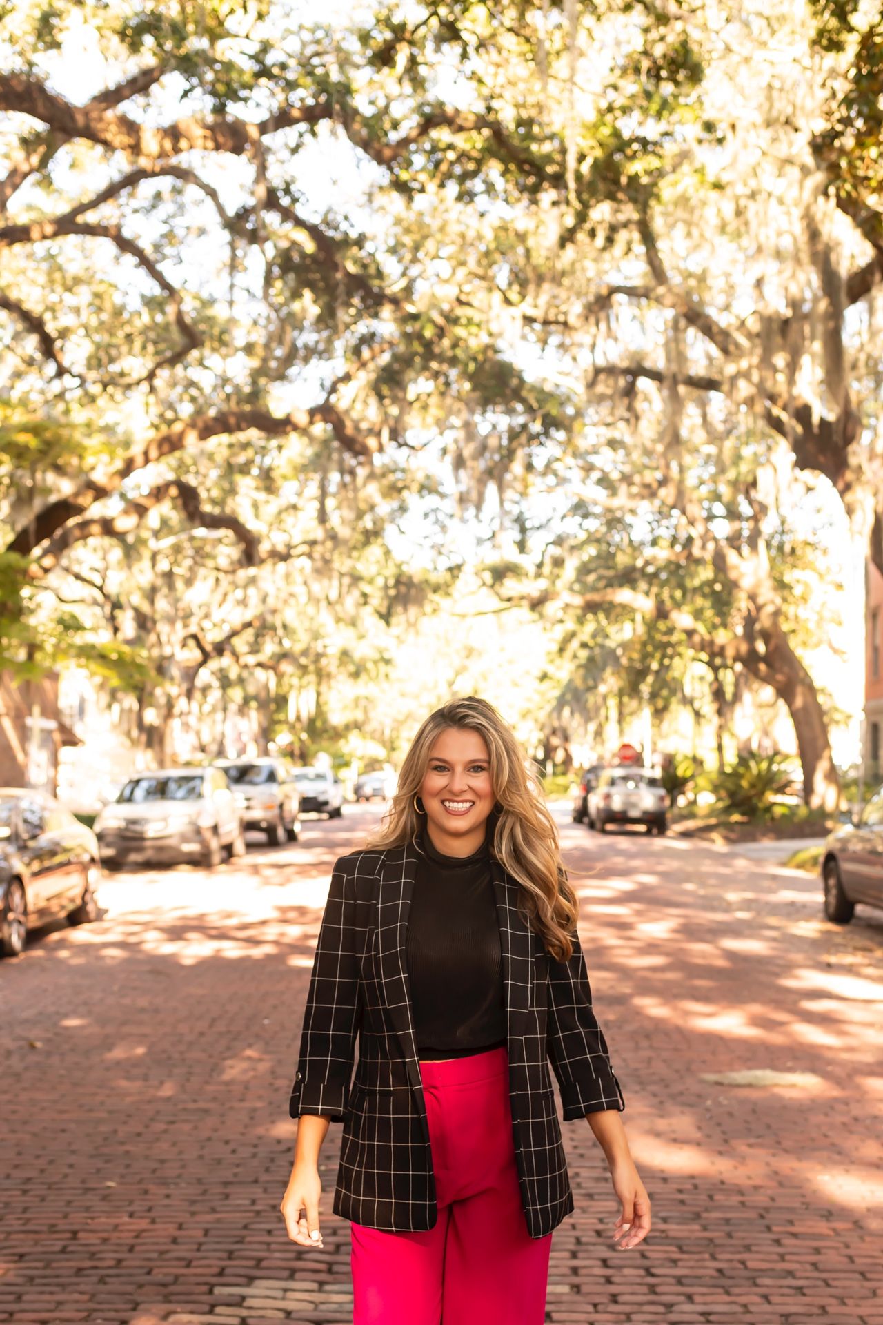 A person wearing a black and white plaid blazer and bright pink skirt stands on a brick path under Spanish moss trees.