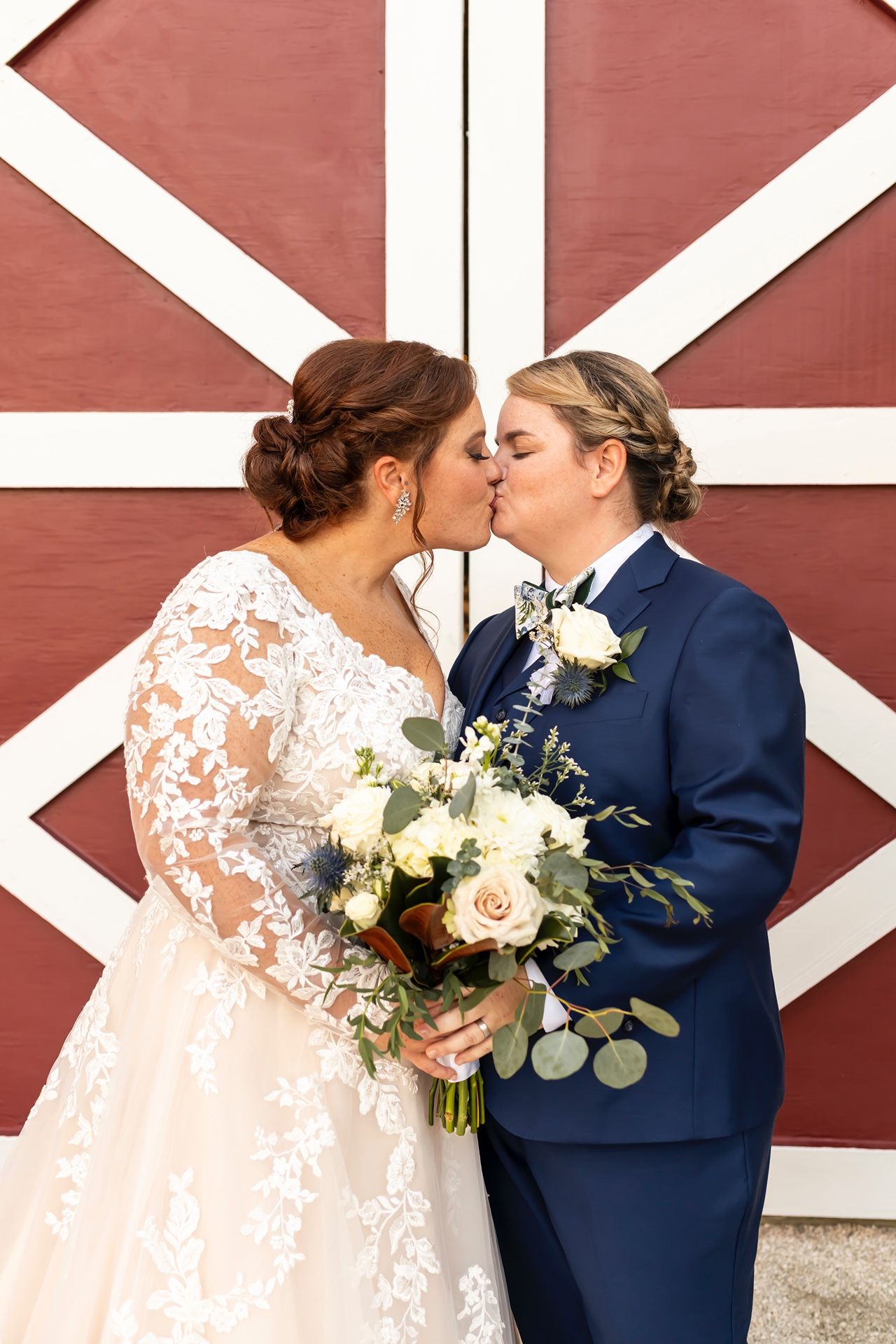 Happy couple sharing a romantic kiss in front of a rustic red and white barn wall while holding a wedding bouquet.