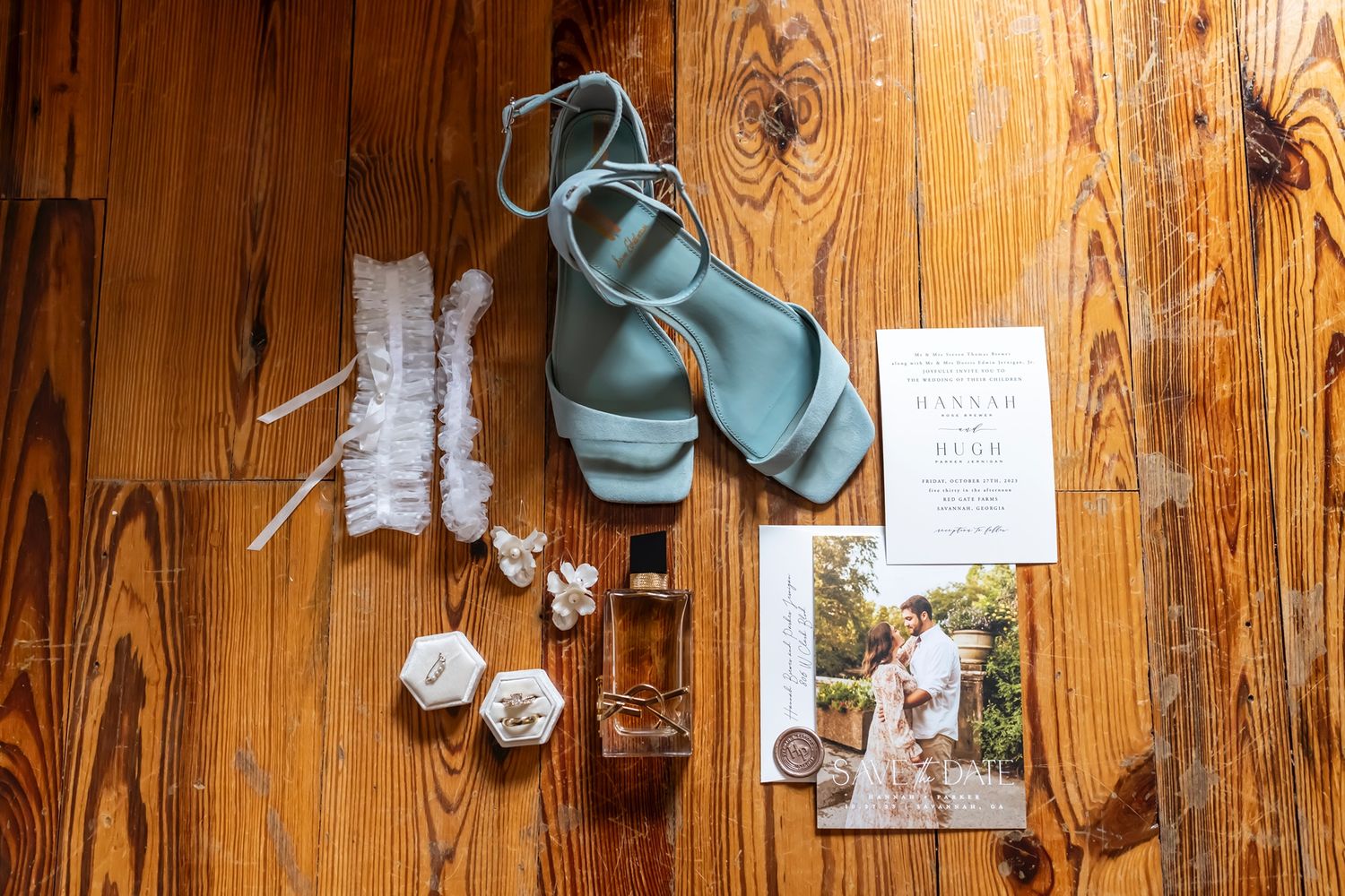 Wedding details including blue heels, garter, rings, perfume and invitation card arranged on wooden floor.