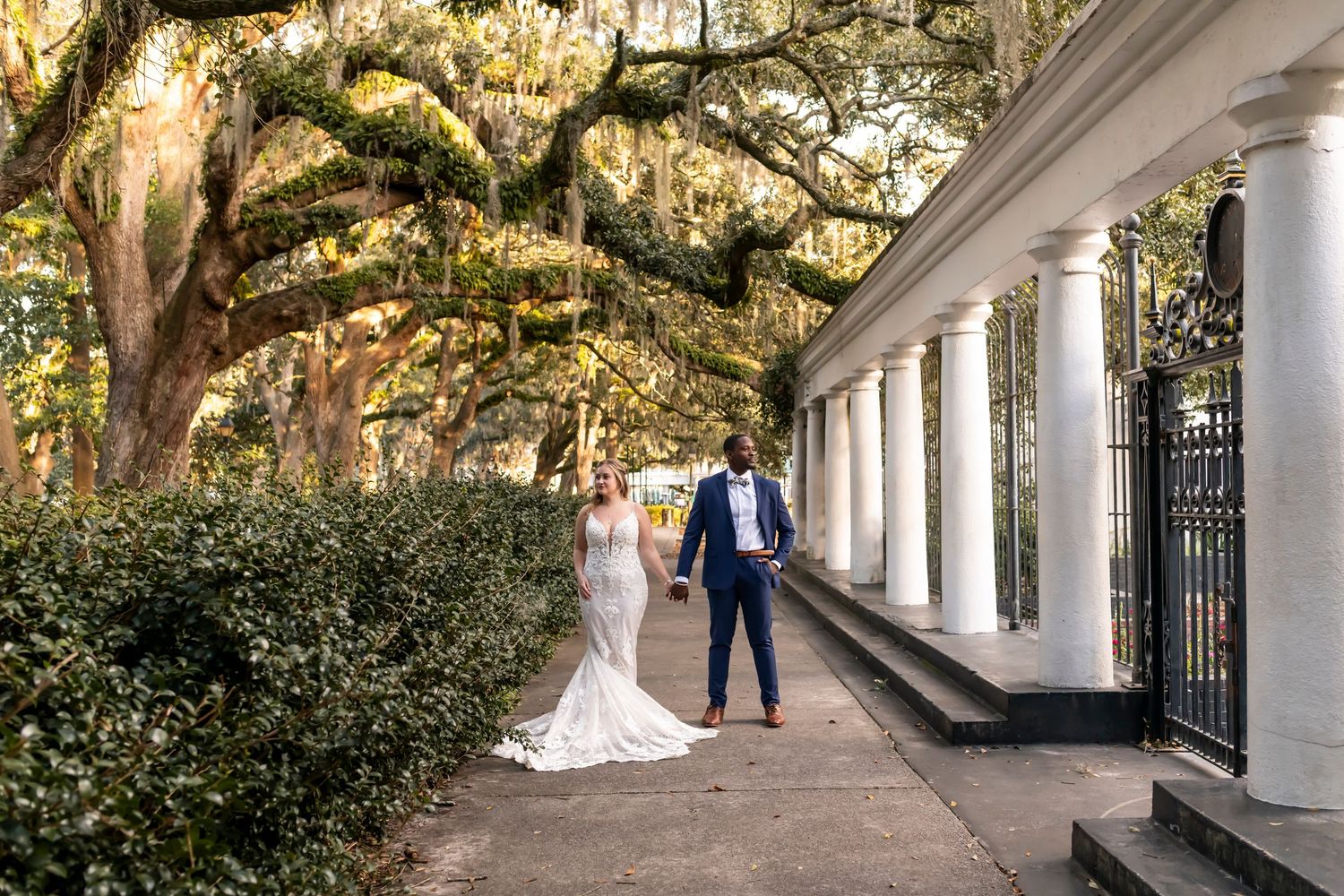Couple stands under grand oak trees draped in Spanish moss along a white columned veranda walkway during golden hour.