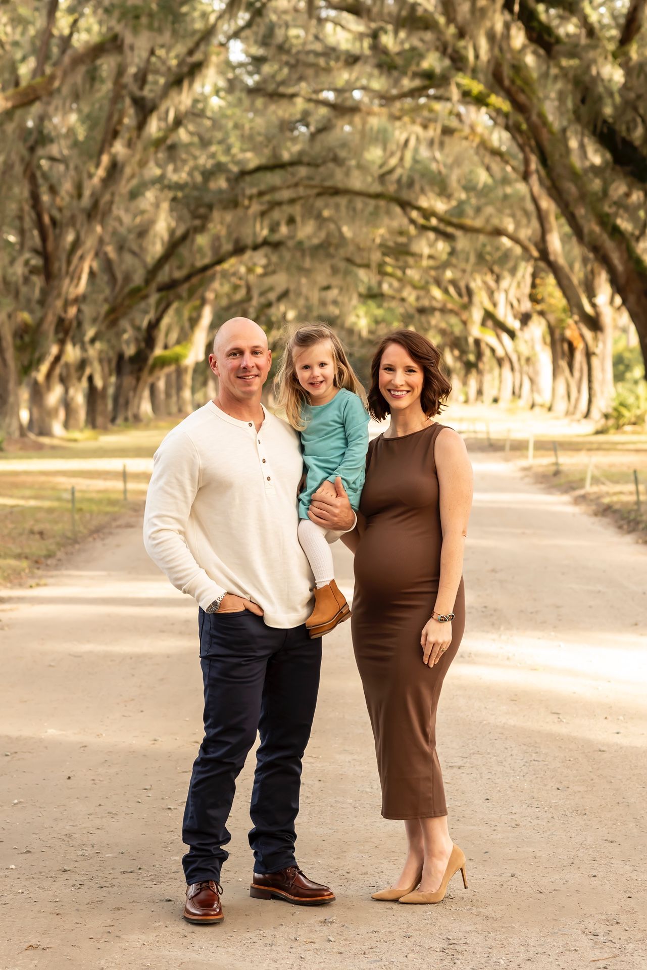 A family poses on a scenic tree-lined path beneath moss-draped oak trees on an autumn afternoon.