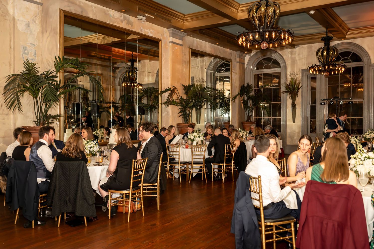 Elegant dinner party in a grand ballroom with chandeliers, mirrored walls, palm plants and guests seated at round tables.