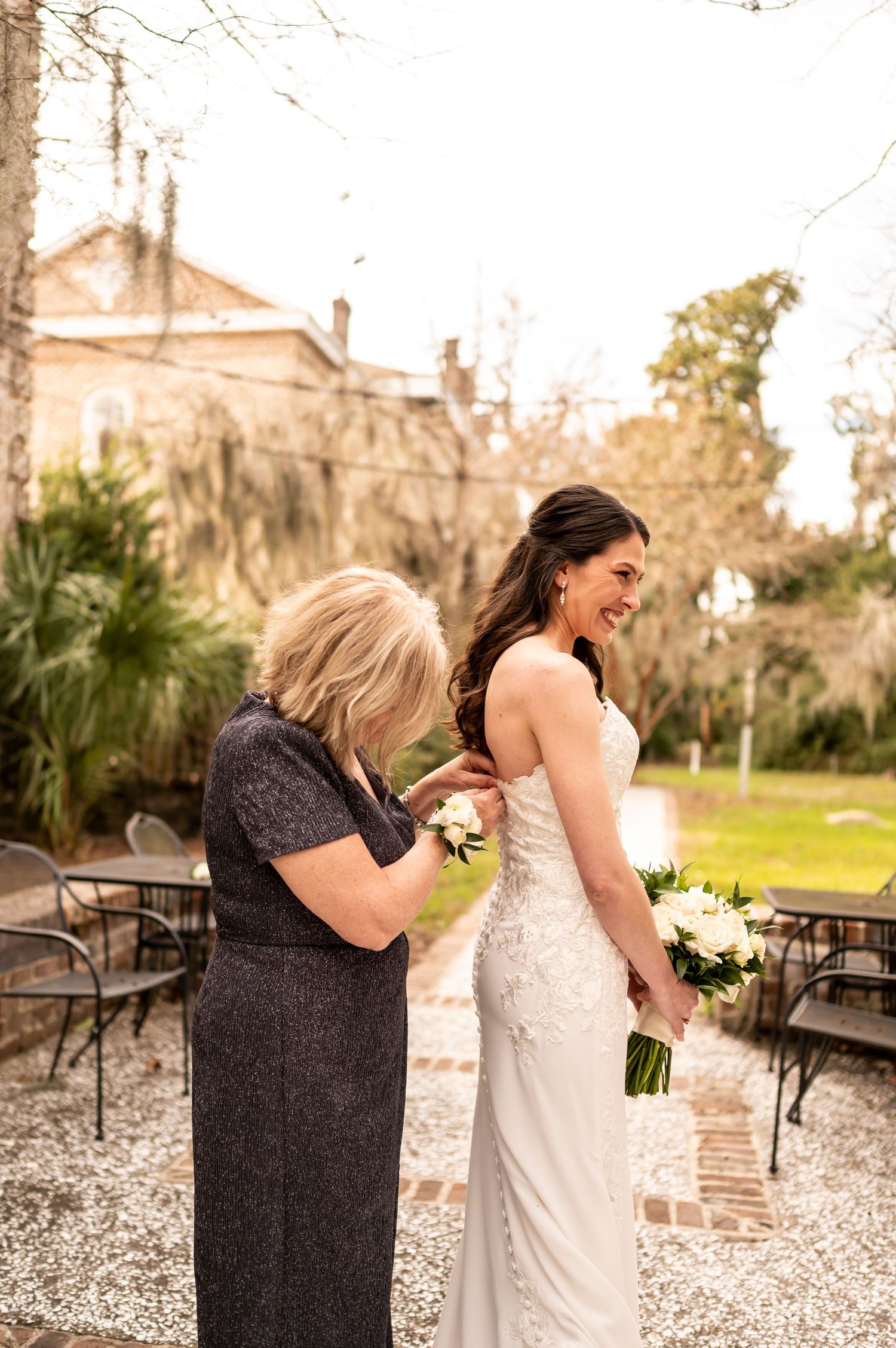 Two people share an emotional moment in formal attire at an outdoor venue with gravel and brick buildings in background.