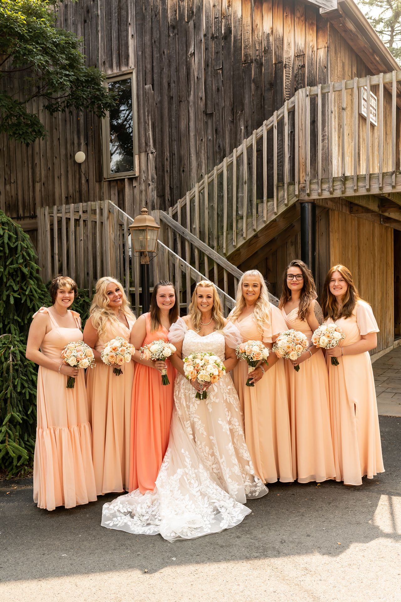 A bridal party in peach-colored dresses poses together against a rustic wooden barn backdrop, holding white bouquets.