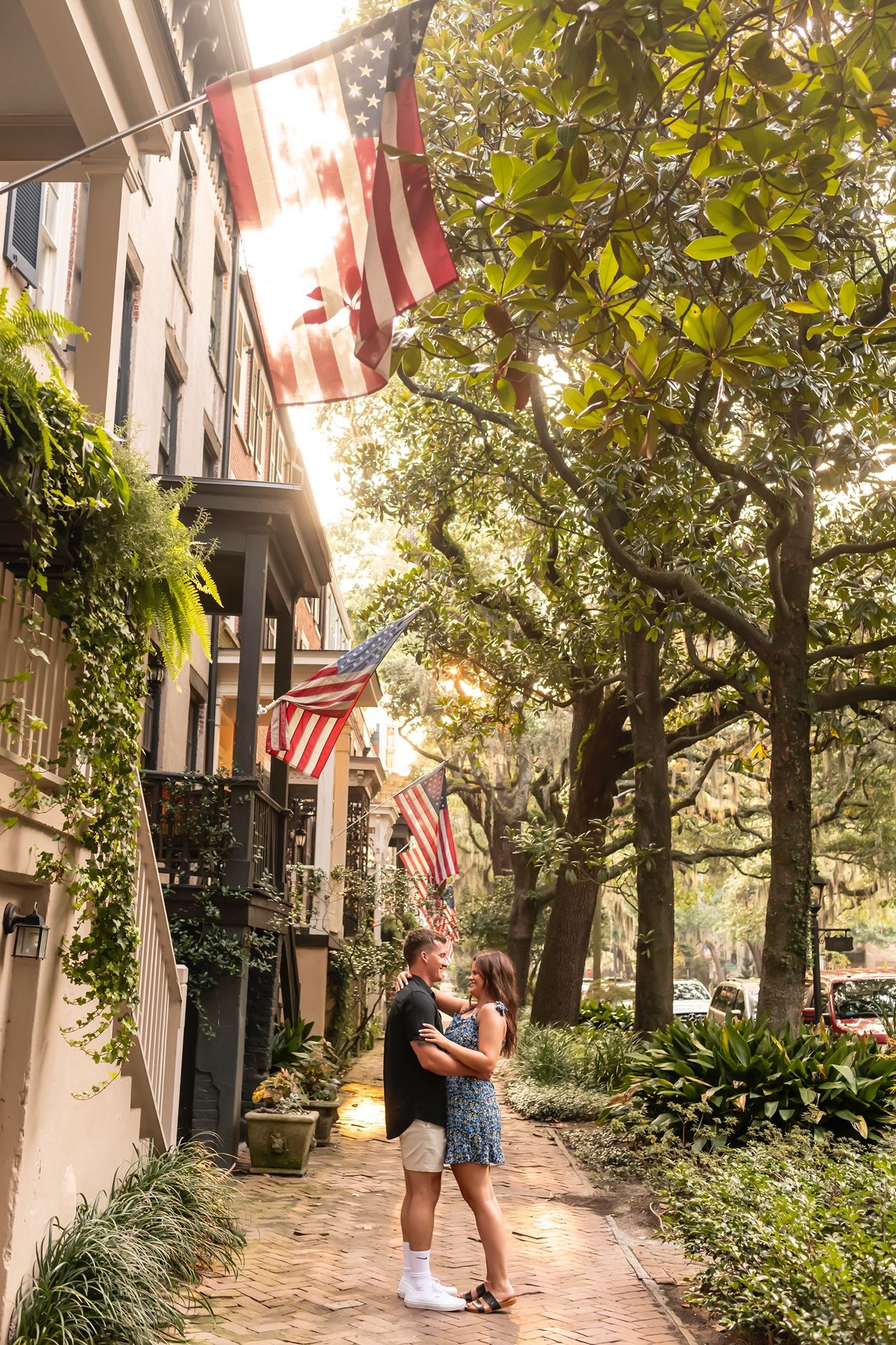 A tree-lined cobblestone sidewalk with American flags displayed on buildings in a historic district.