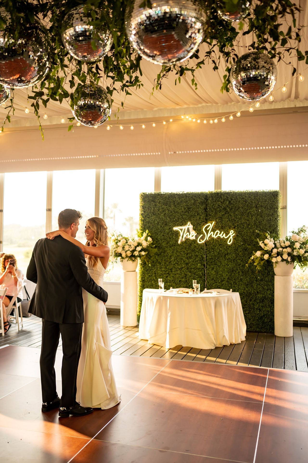 Couple shares first dance at elegant wedding reception with greenery wall backdrop and hanging disco balls on display.