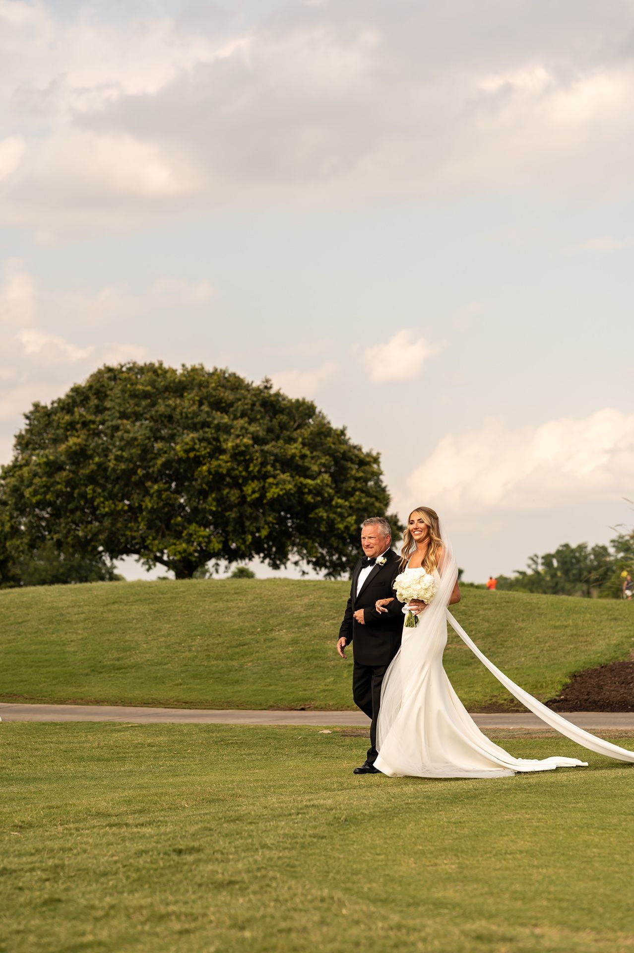A couple sits together on a grassy hill during sunset with a long flowing wedding veil trailing behind them.