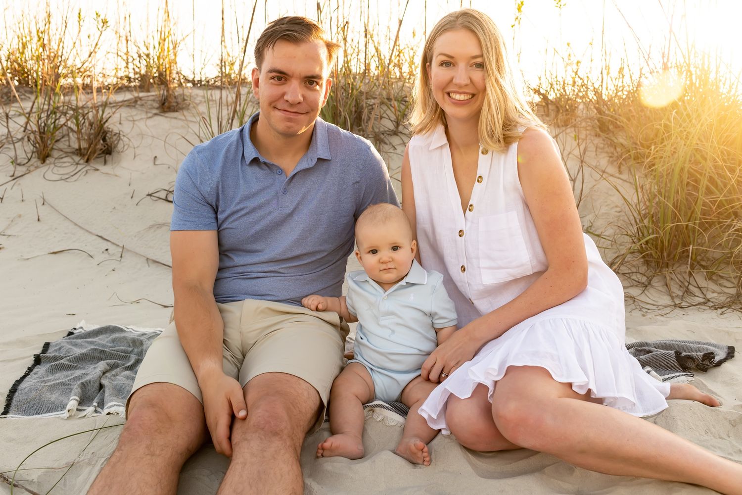Family sits together on beach blanket at sunset with sand dunes and golden light in background.