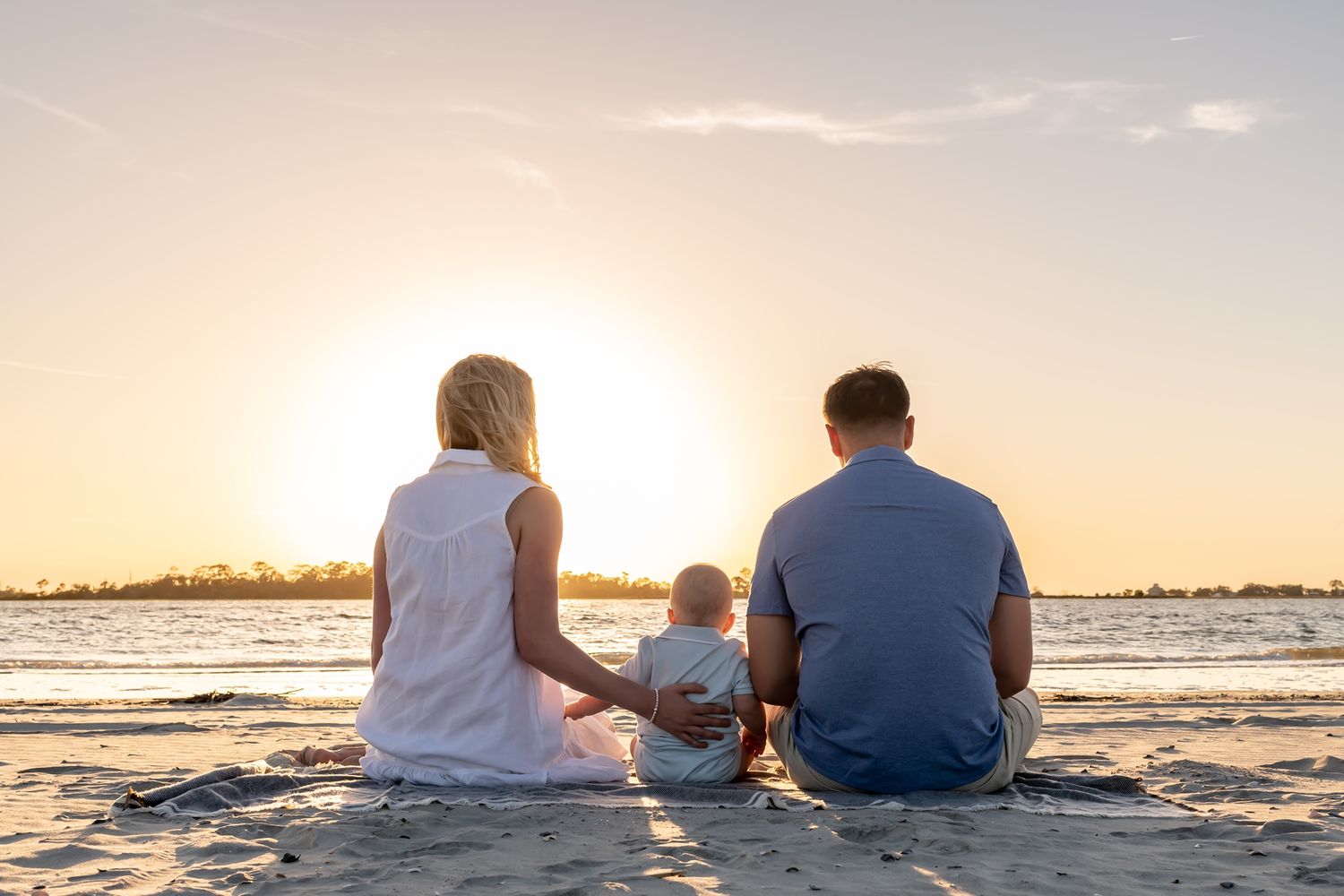 Family of three sits together on sandy beach watching sunset over ocean horizon on peaceful evening.
