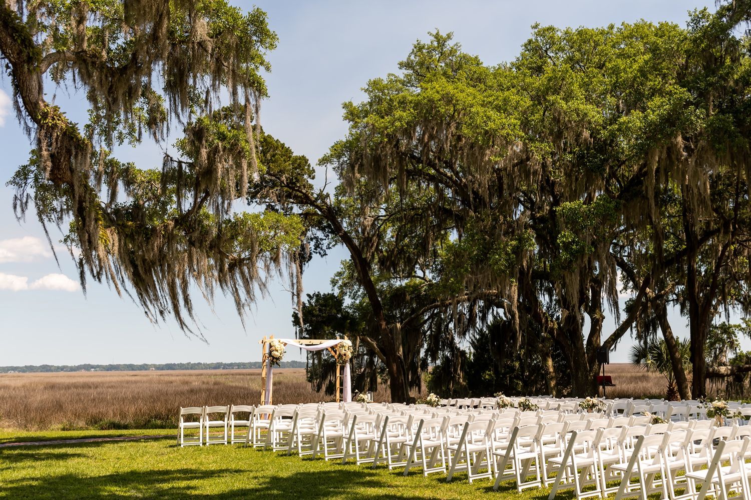 White chairs set up for an outdoor ceremony under Spanish moss-draped oak trees overlooking coastal marshland.