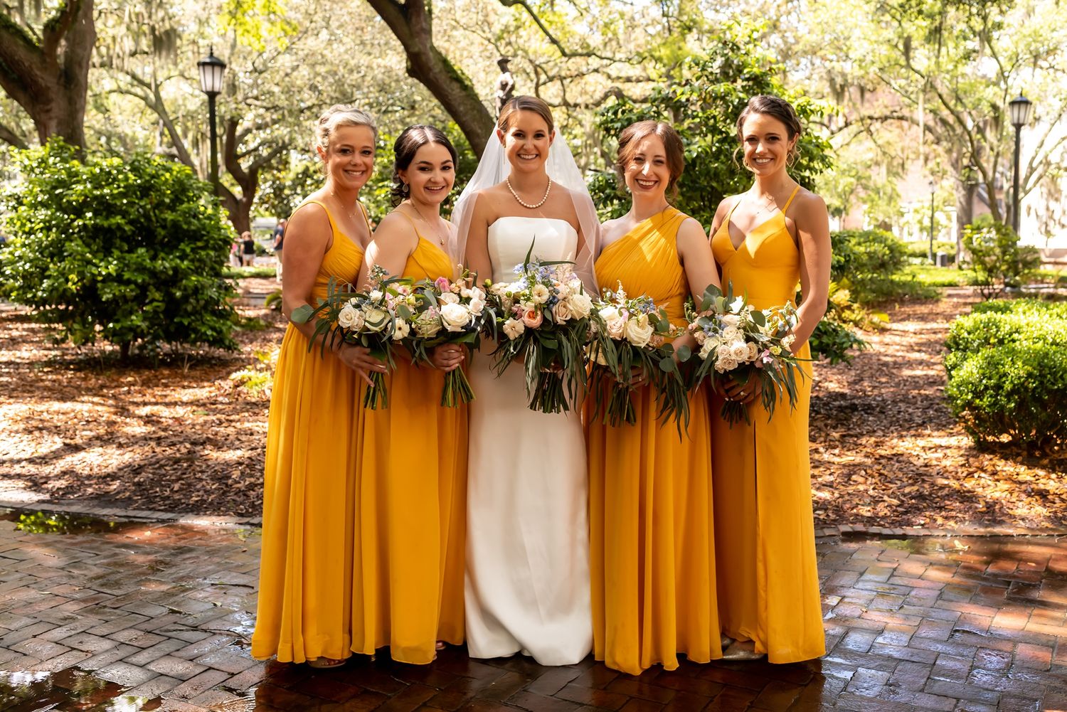 A bride in white stands with bridesmaids wearing marigold yellow dresses while holding floral bouquets in a garden setting.