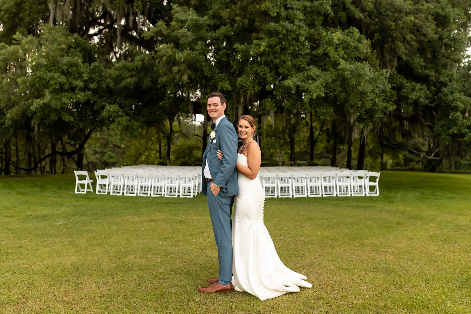 A couple stands on a lush green lawn with rows of white chairs set up for an outdoor wedding ceremony.