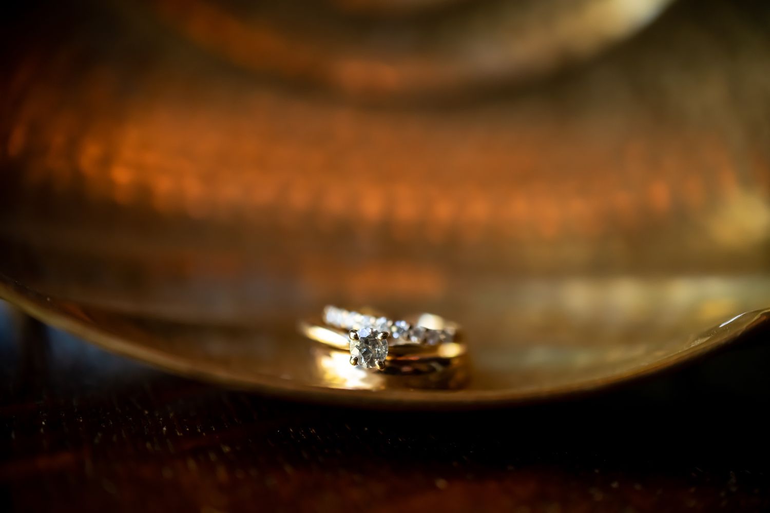 A sparkling engagement ring sits in soft focus on a reflective surface with warm ambient lighting.