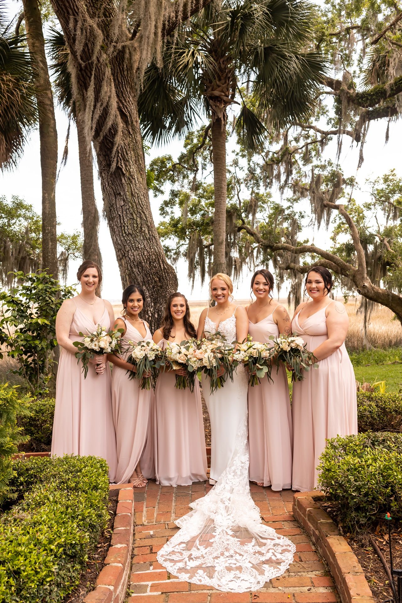 Wedding party in blush pink dresses poses with bouquets on a brick pathway under Spanish moss draped trees.