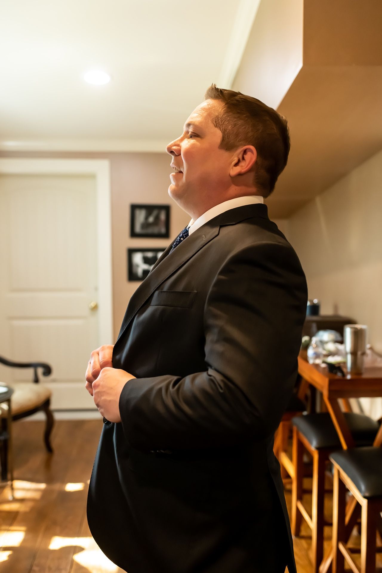 A person in a black suit jacket adjusts their attire while standing in a warmly lit room.