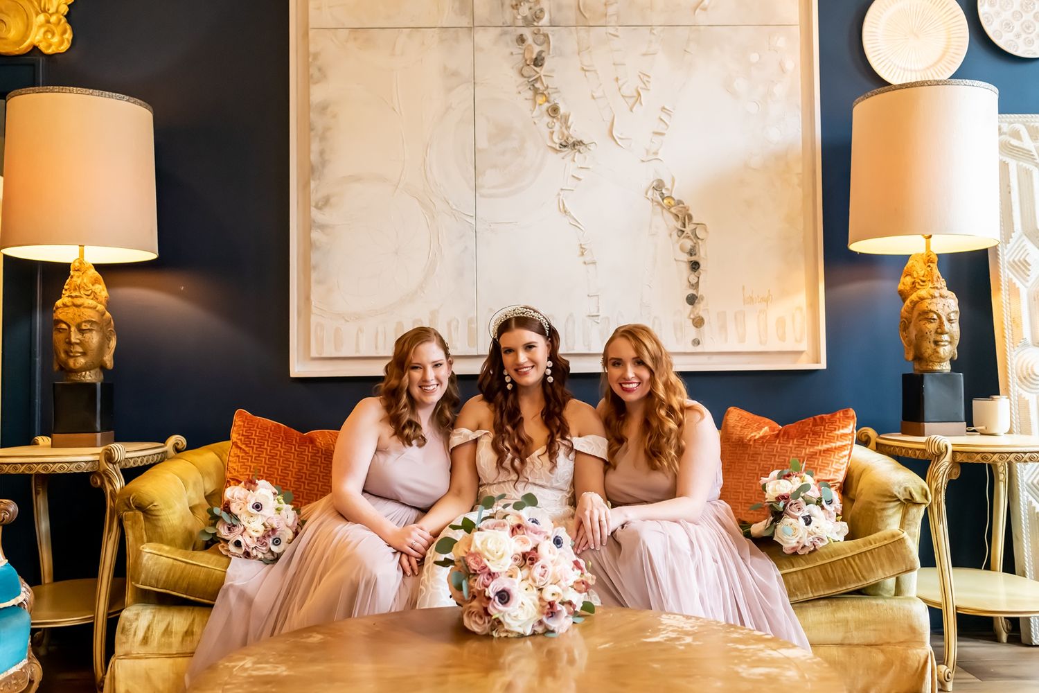Three bridesmaids in pink dresses sit together on a gold couch with bouquets in an elegant room with gold lamps.