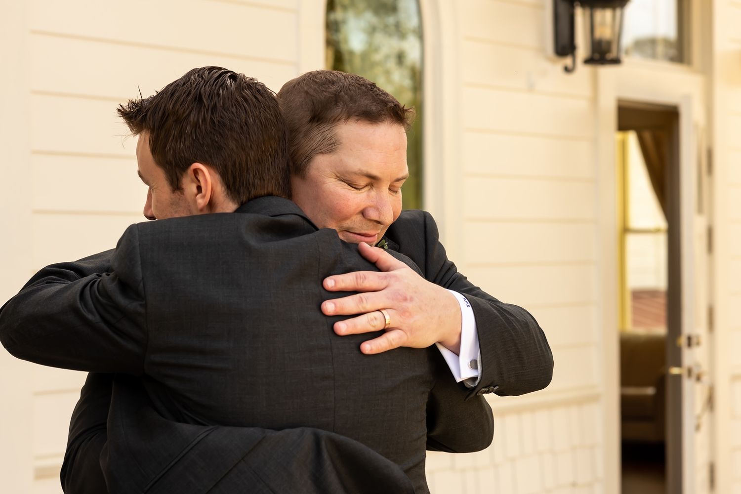 Two people in formal attire share a heartfelt embrace outside a building with white siding and black trim.
