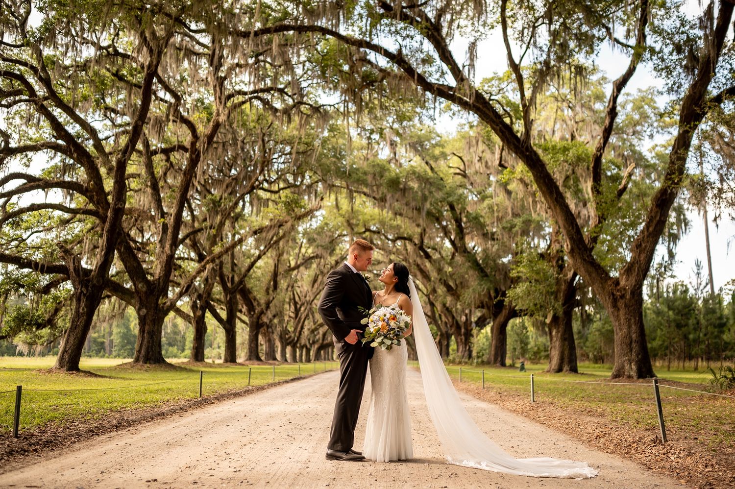 A couple stands together on a dirt road lined with oak trees covered in Spanish moss.