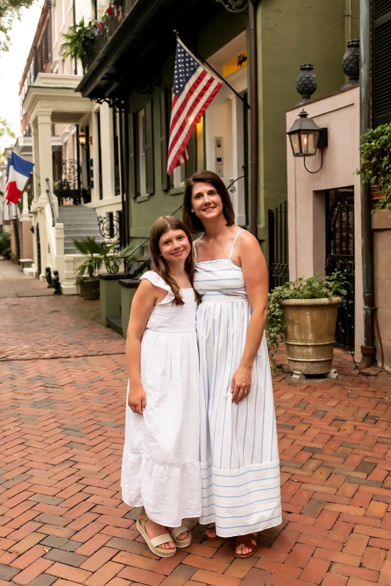Two people in white dresses pose together on a historic brick street lined with colonial buildings and American flags.