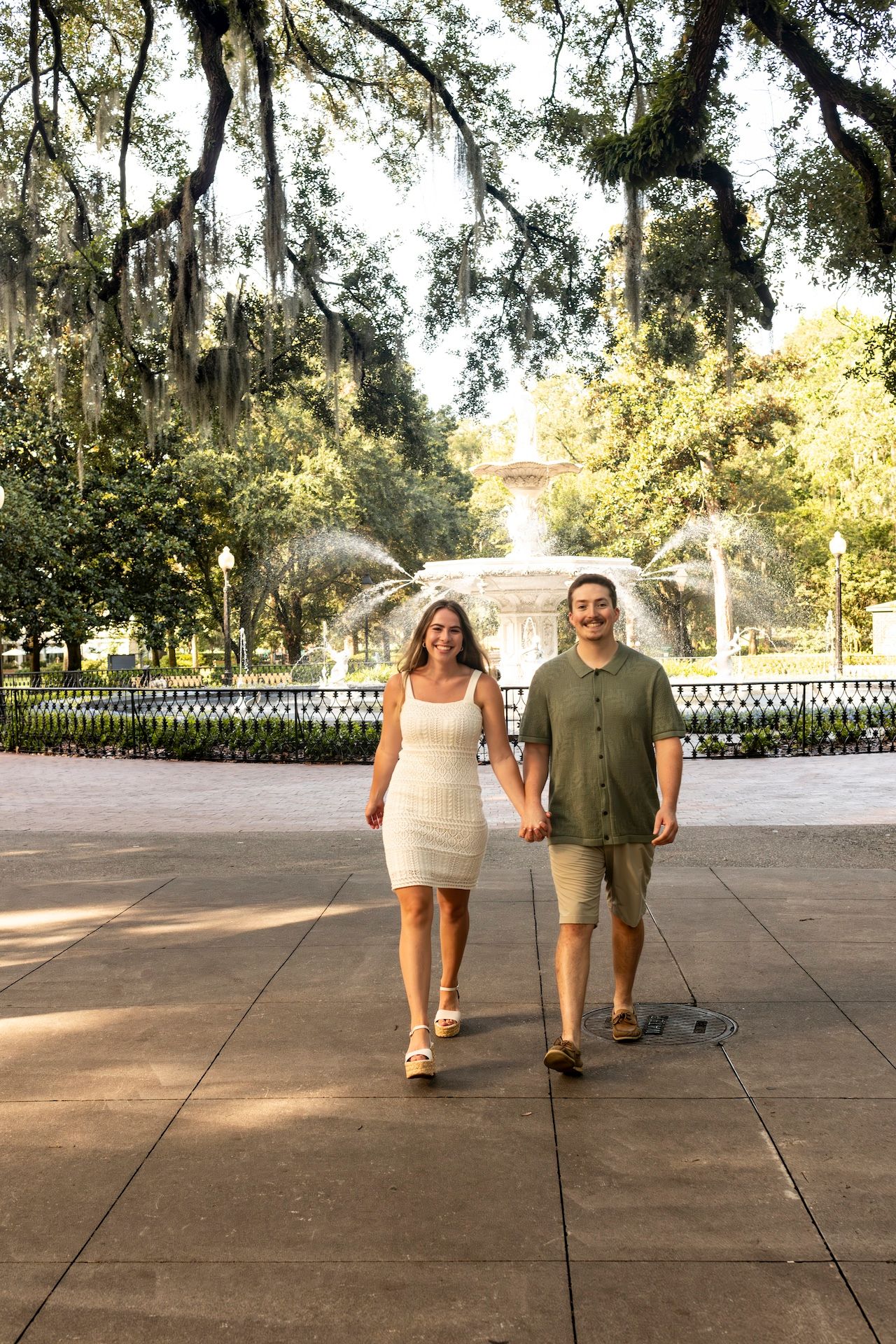Two people in casual attire walking together on a paved walkway surrounded by trees and a white fence on a sunny day.