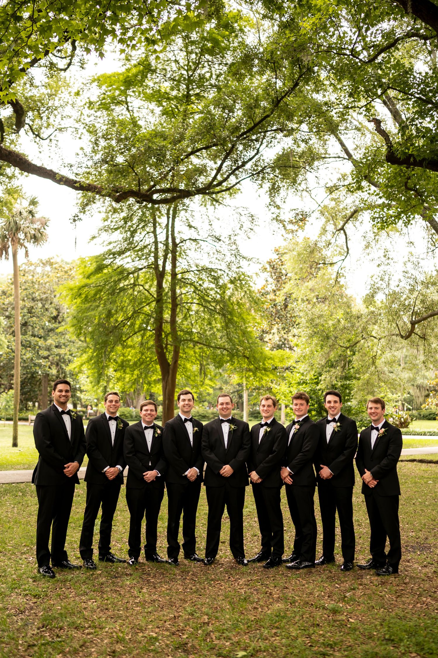 A line of groomsmen in black tuxedos pose together under green trees in an outdoor setting.