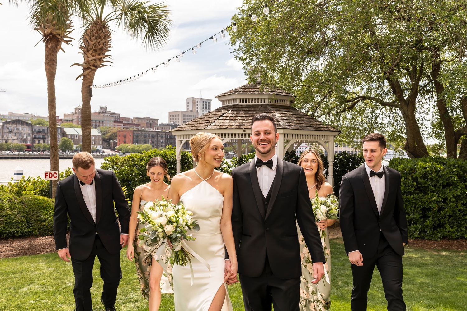 Wedding party dressed in black and white formal attire poses together outdoors with palm trees and greenery in background.