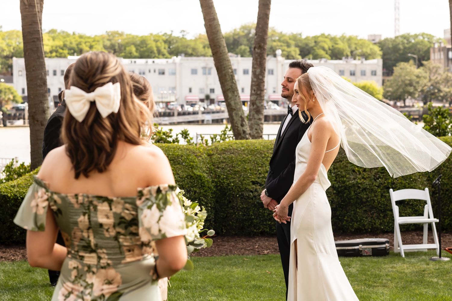 Wedding party celebrates outdoors on a sunny lawn with white folding chairs and decorative floral arrangements.