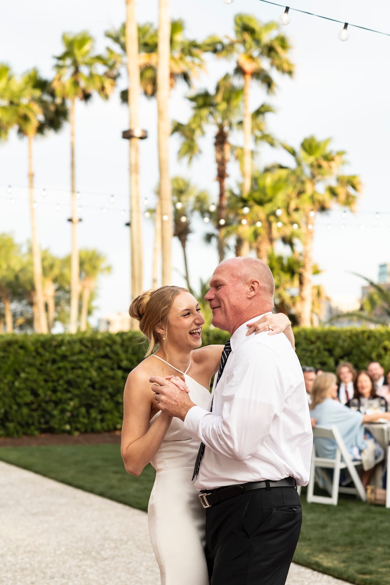 A joyful dance moment between a bride in white dress and her dance partner amidst palm trees at an outdoor reception.