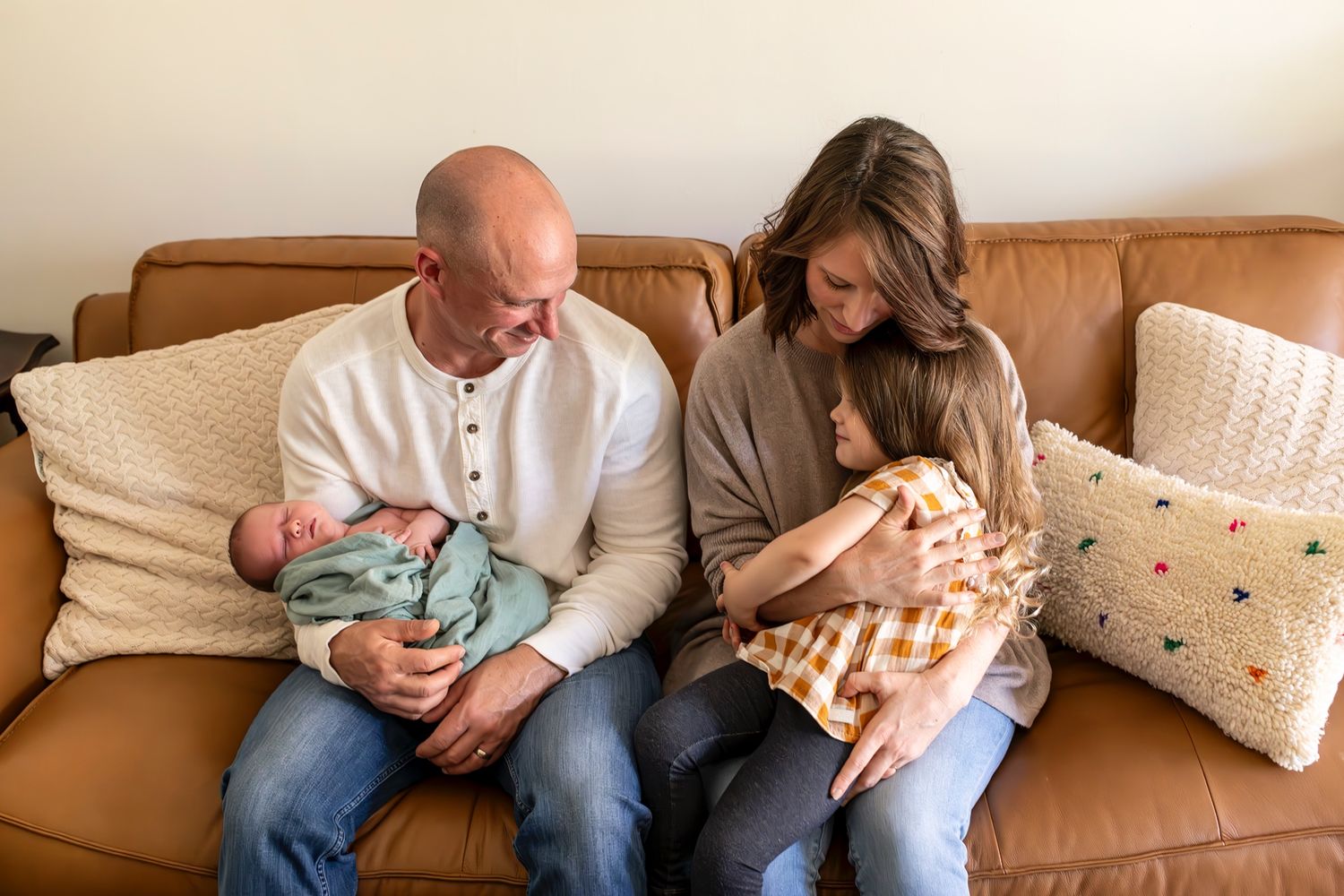 Two people sitting on brown leather couch holding newborn babies while surrounded by decorative throw pillows.