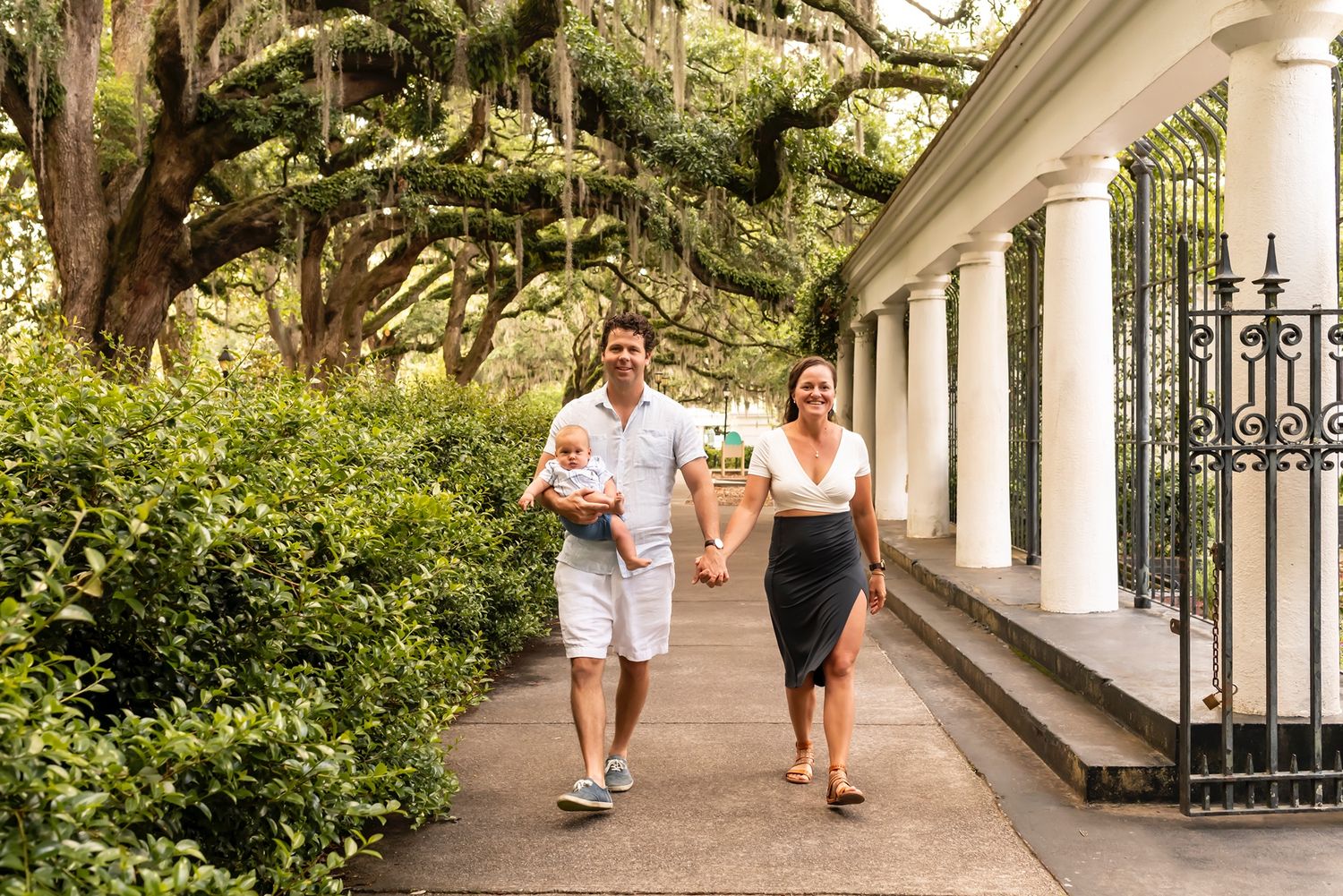 A couple strolls along a historic colonnade walkway beneath Spanish moss-draped oak trees in the American South.